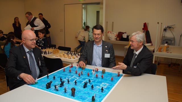 Premiere in der Geschichte des Makruk: An der Universität Hamburg wurde im Mai 2012 zum ersten Mal ein Turnier im Siamschach außerhalb der Grenzen Thailands veranstaltet. Zur Eröffnung ließen sich an einem Demonstrationsbrett, das Turnierorganisator René Gralla gebastelt hatte (re.), der Dekan des Fachbereichs Thaiistik, Professor Volker Grabowsky (li.), und Stefan Krohn als Vertreter des Königlich Thailändischen Honorargeneralkonsulats (in der Mitte) das besondere Szenario des Makruk erklären. Der Clou: das Spielfeld ist blau eingefärbt, weil es ein Überflutungsgebiet symbolisiert; schließlich kommen im Thaischach auch Boote zum Einsatz (im Bild jeweils in den Ecken des 64-Felder-Plans postiert). Foto: Jeerathon Srongsuwan